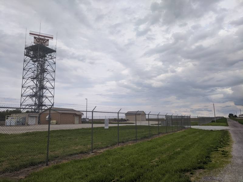 A radio tower is in a fenced off area. There are a couple of small buildings nearby. Photo by Tom Ackerman.