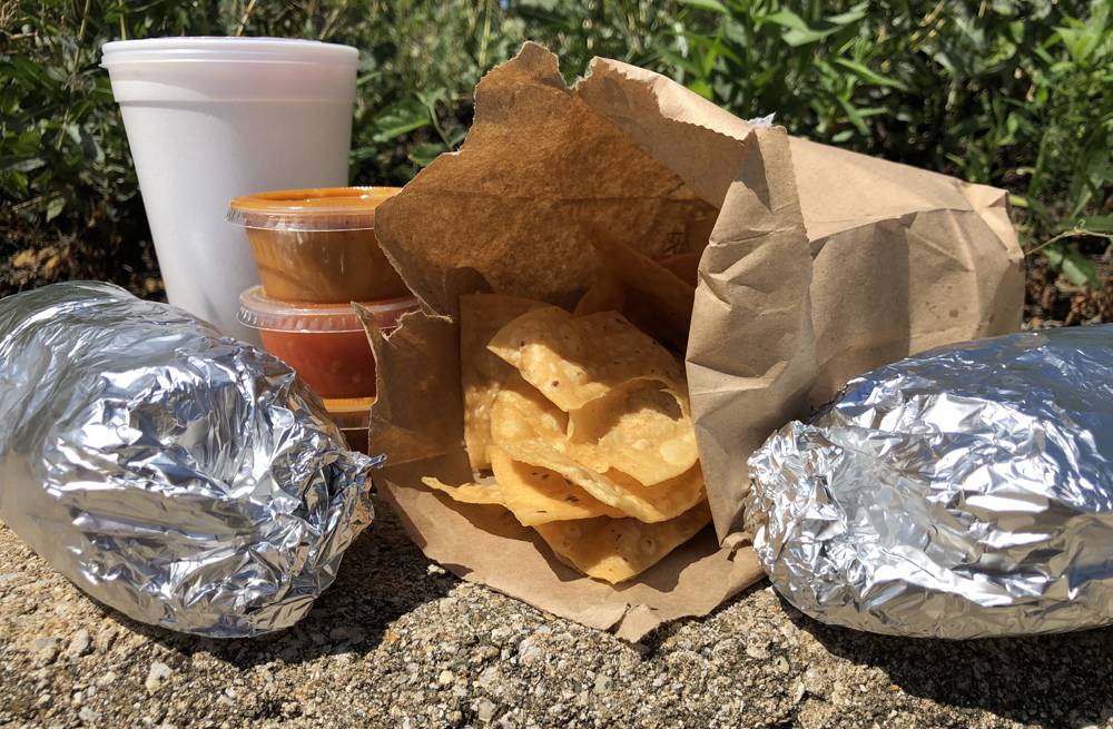 A to go drink in a white styrofoam cup with a cover, a stack of three tiny sauce cups with covers, two tin-foiled wrapped burritos, and an open bag of tortilla chips sits on a concrete bench outside with bushes behind. Photo by Jessica Hammie.