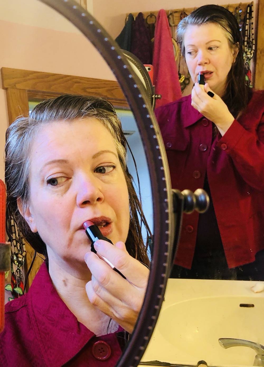 A woman looks into a large bathroom vanity mirror. She is putting on lipstick. The reflection also appears in a smaller, round countertop mirror. Photo by Danielle Chynoweth. 