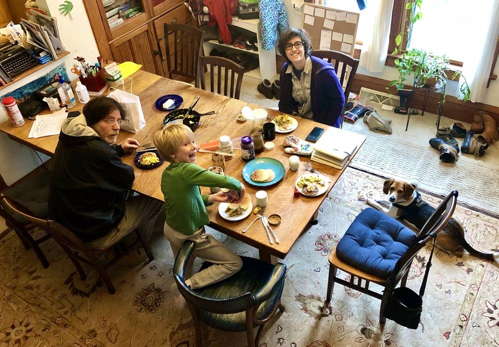 Two adults and a child are at a long dining table. There are breakfast dishes on the table. There is a beagle laying on the floor nearby. The view is above, likely from a staircase overlooking the dining area. Photo by Danielle Chynoweth. 