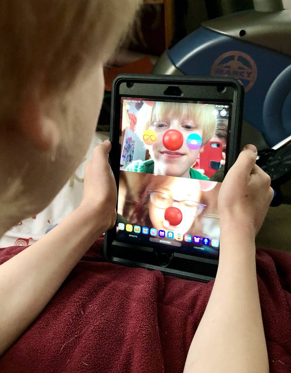 A child holds a tablet and is video chatting with a woman. On the tablet screen, both faces are covered in clown facial markings, including big, red noses. Photo by Danielle Chynoweth. 