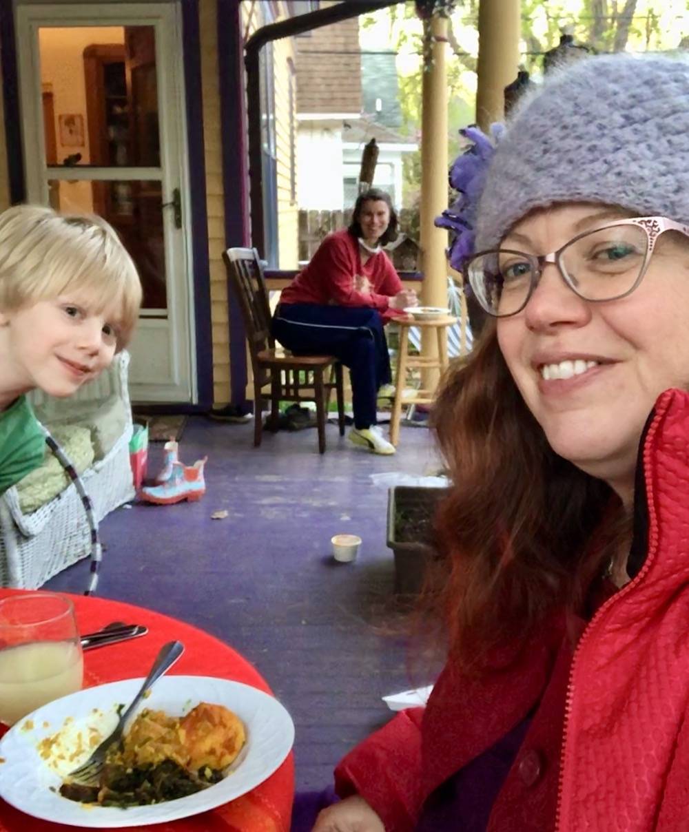 On a large front porch, Danielle Chynoweth is pictured in the foreground, wearing a knit hat and a red jacket. There are bowls of food on the table in front of her. Her son Ezra is pictured on the left, leaning into the frame. On the other end of the porch is Miriam Larson, who is seated alone and also eating. Photo by Danielle Chynoweth. 
