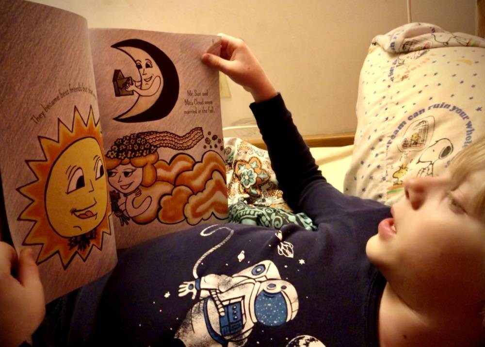 A boy reads an illustrated book while lying in bed. He is wearing a blue astronaut shirt. Photo by Danielle Chynoweth. 