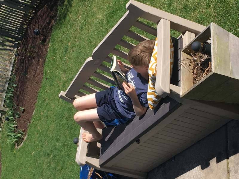 A boy is stretched out on a bench on a backyard patio. He is reading a book. Photo by Erin Ewoldt. 