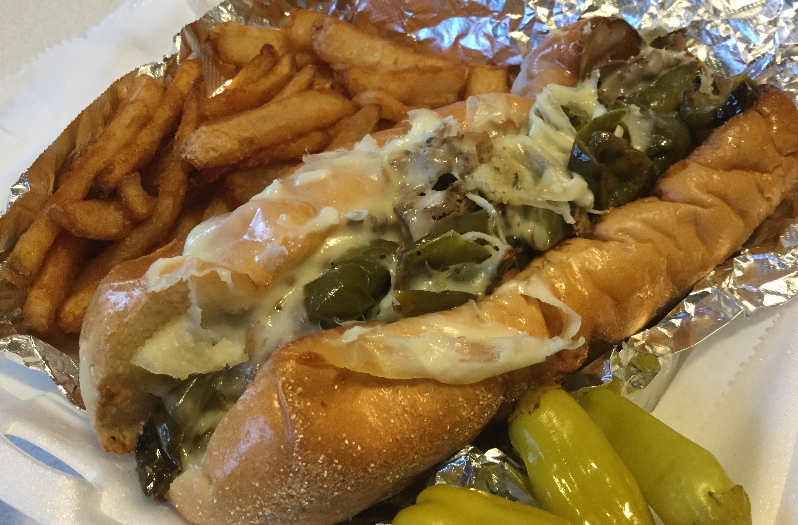 An italian beef sandwich sits on tin foil in a white styrofoam carryout container with fries above the sandwich and several banana peppers below it. Photo by Rachael McMillan.