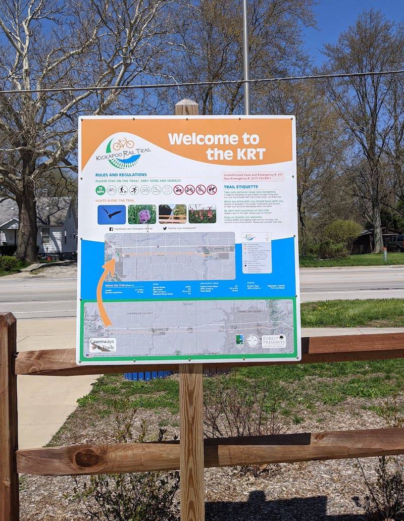 An orange, blue, and white sign announcing the entrance of the Kickapoo Rail Trail. It sits on a wooden fence. Photo by Katriena Knights.