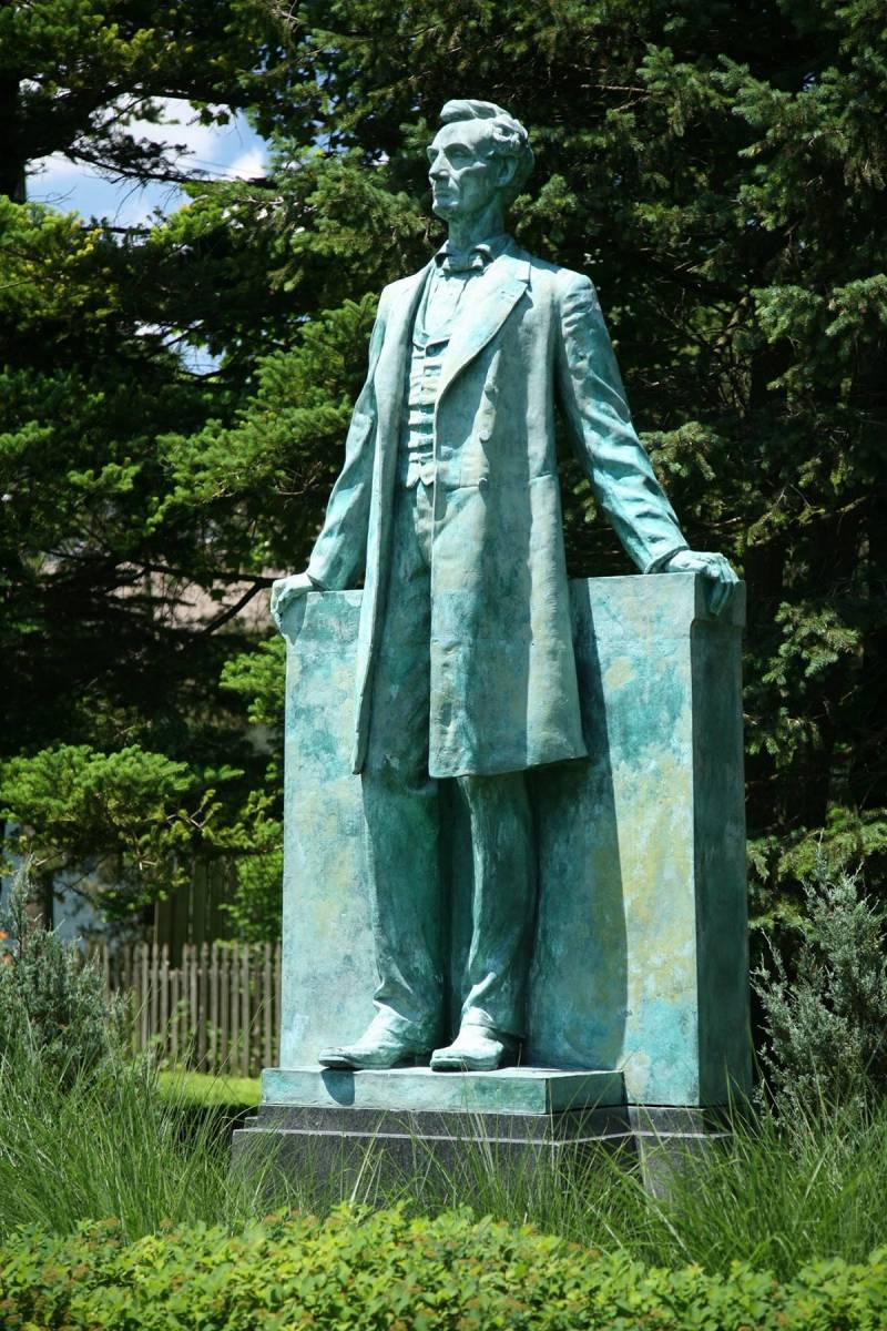 Bronze Lincoln Statue, slightly larger than life, standing on a small bronze platform atop a 3' high granite pedestal.  Lincoln leans slightly back with outstretched arms resting on a bronze backpiece roughly waist high.