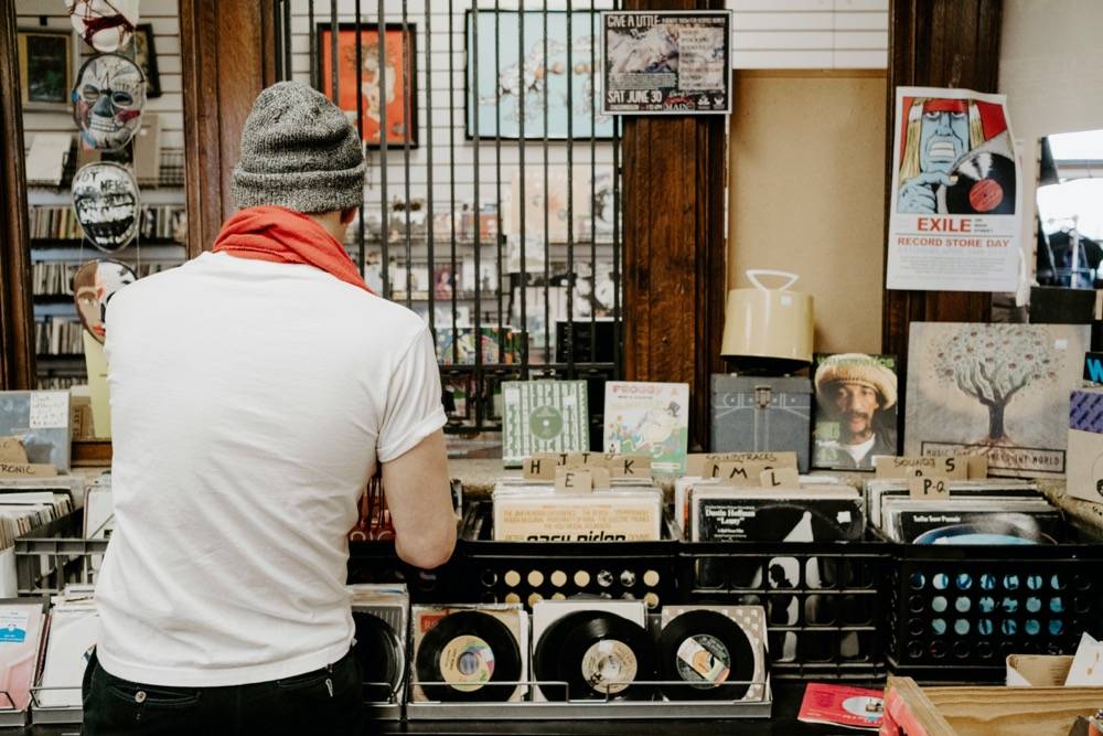 IMAGE: View of man, from behind, as he stands looking through a box of records. The man wears a white shirt, red scarf, and grey stocking cap.  Photo by Anna Longworth.