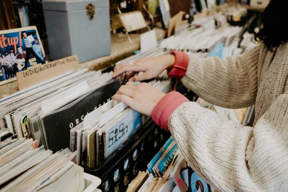 IMAGE: View of two hands touching a box of records amongst a row of many boxes. The woman wears a white sweater with red cuffs. Photo by Anna Longworth.