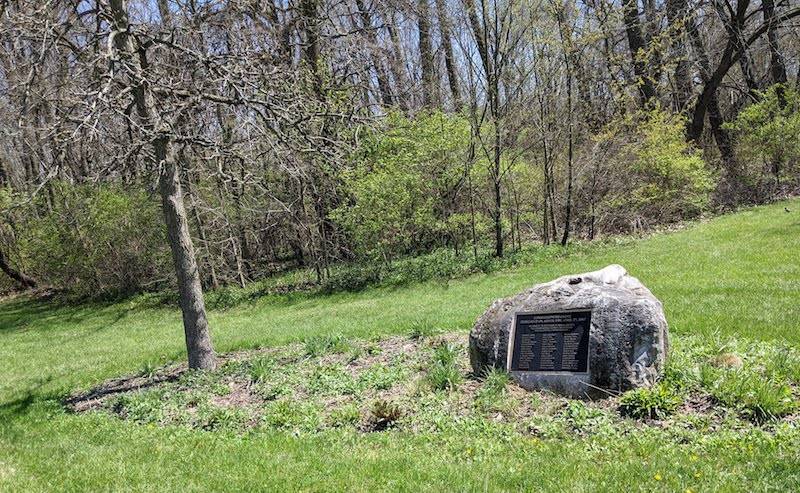 A stone marker sits in a grassy area adjacent to a small tree. There is a metal plaque in the center of the stone. Photo by Katriena Knights.