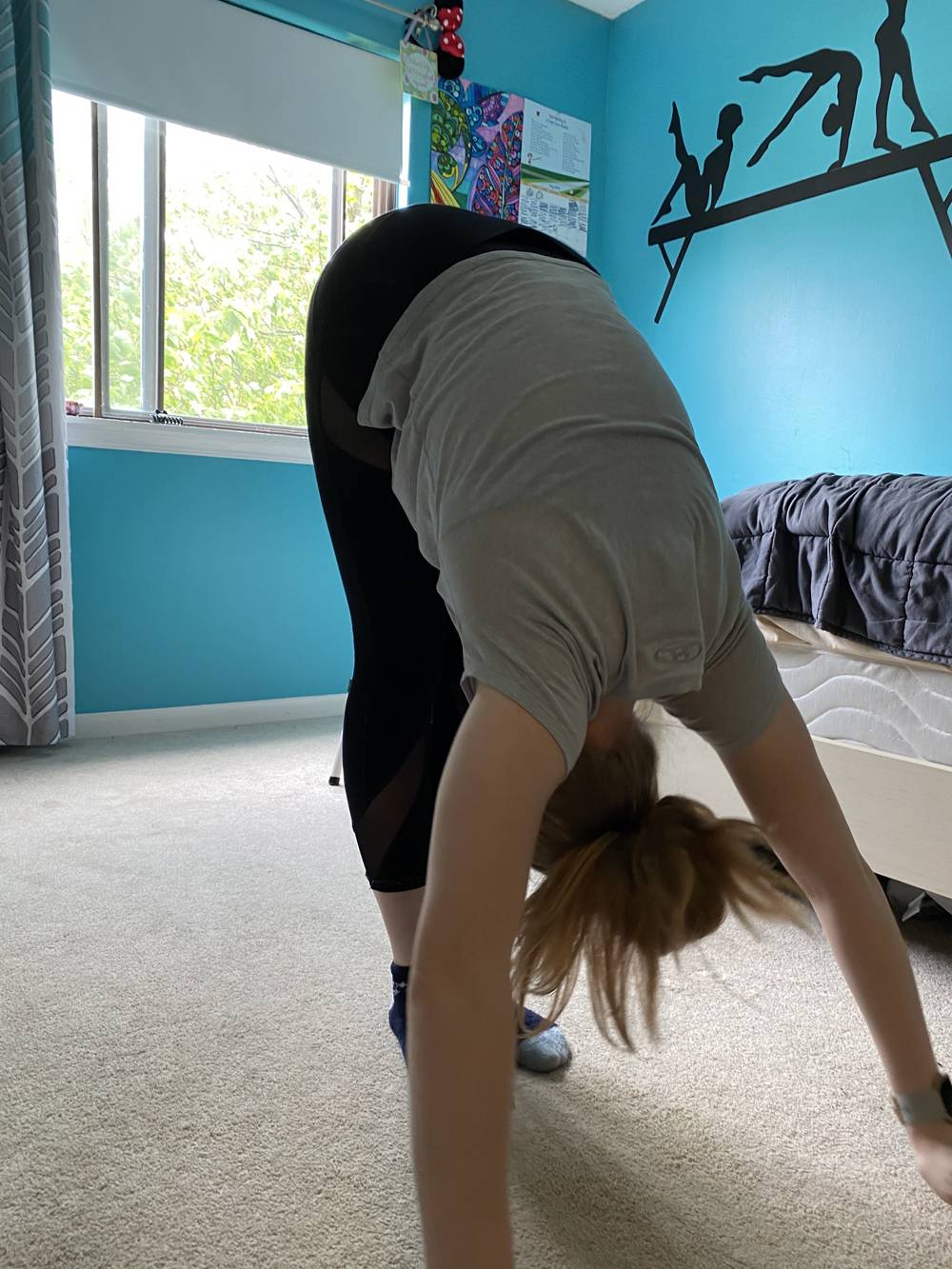 A teenage girl is bending forward at the hips, with hands pressed to the floor. Her blonde ponytail dangles as her body bends forward. She is in a bedroom that is painted bright blue. Photo by Elena Reifsteck. 