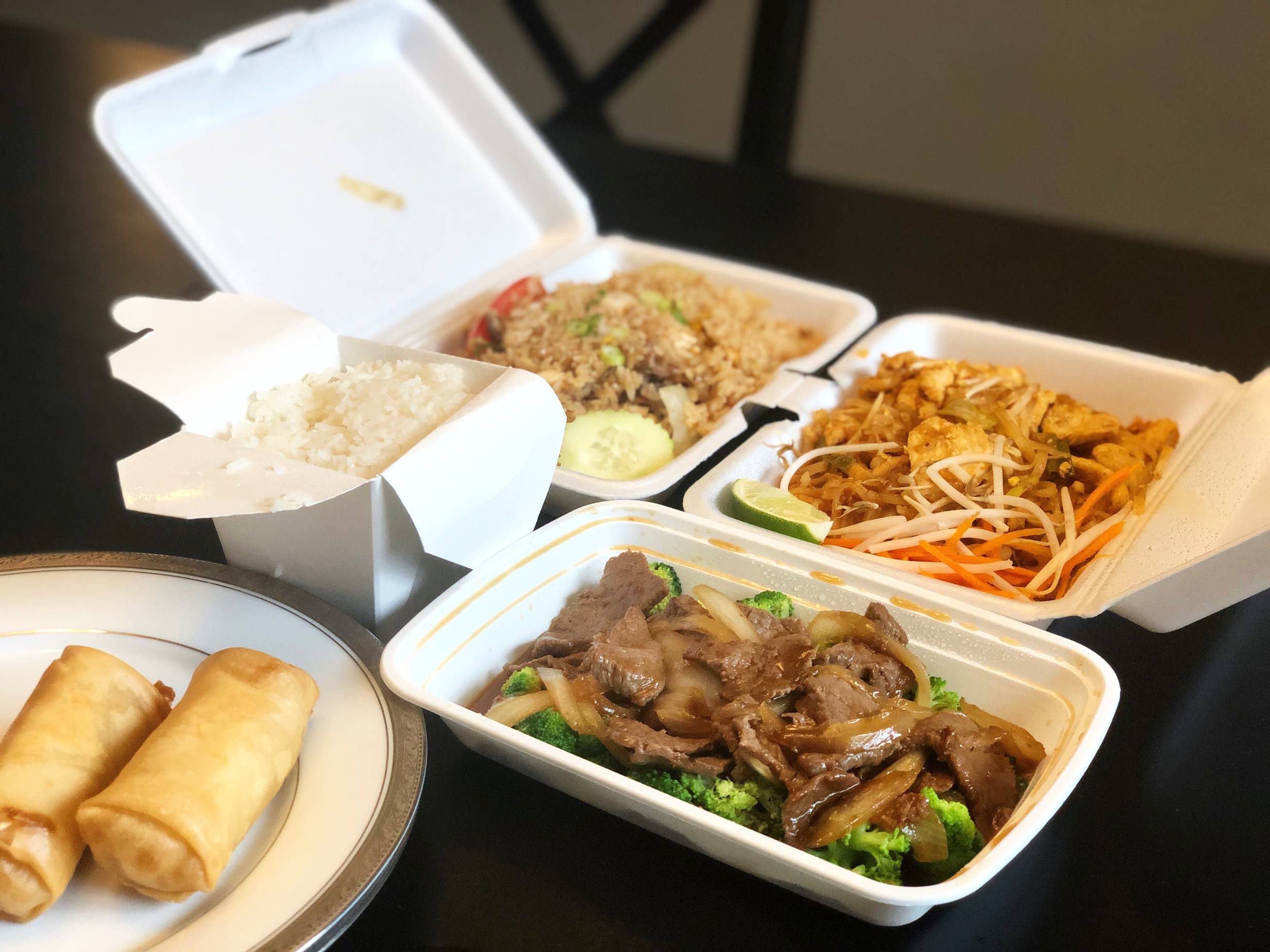 All the takeout options for the author's dinner. Two egg rolls on a plate, a carton of steamed white rice, a plastic container of beef and broccoli, a container of pad thai, and a container of fried rice on a black table. Photo by Alyssa Buckley.