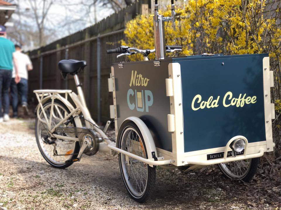 a cart with taps is attached to a bike that acts as NitroCup's mobile serving station. It sits in front of a fence and bush. Photo by Nitrocup Facebook.
