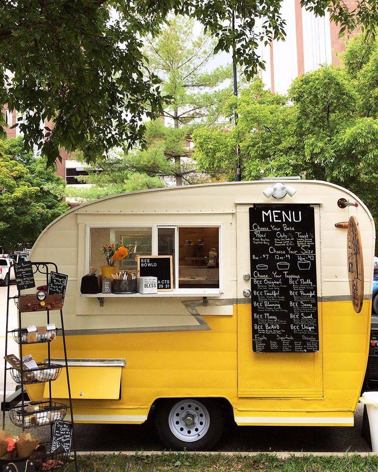 Just Bee's vintage yellow and cream Shasta parked by a curb amongst trees. Displayed are its chalkboard menu and a wire rack to the side that is full of various local products for sale. Photo by Just Bee Acai.