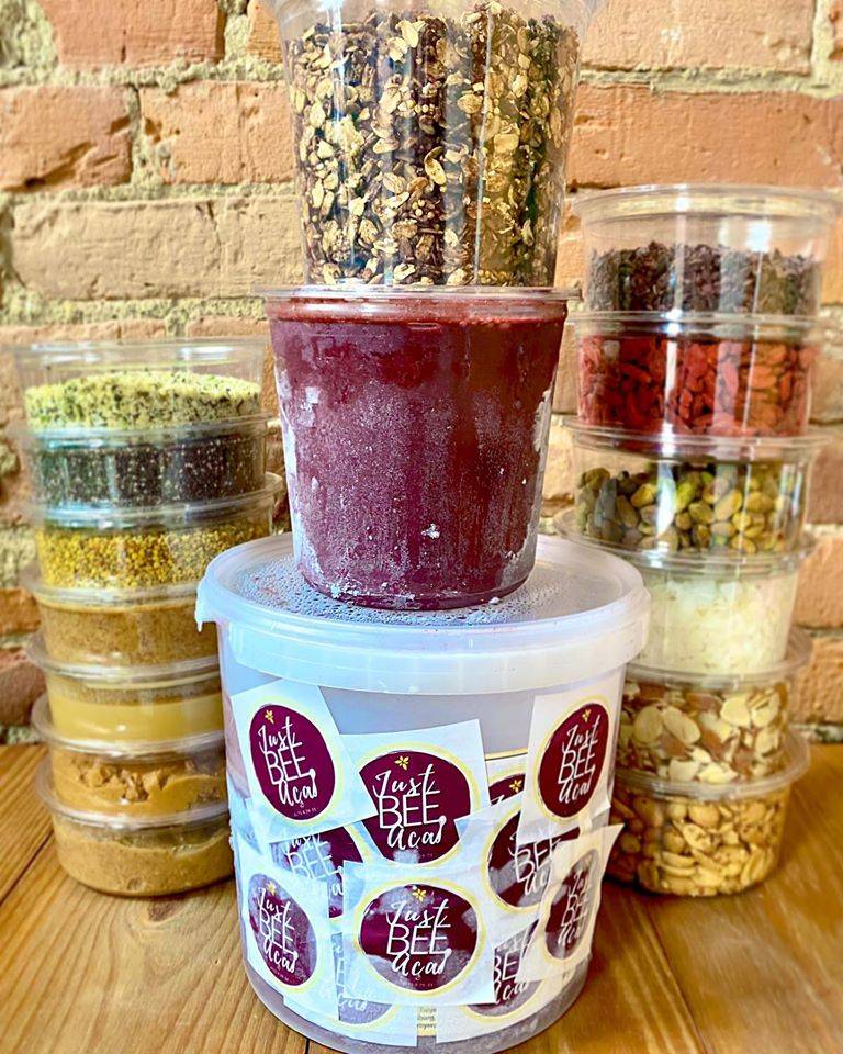 Tubs of various aÃ§aÃ­ bowl ingredients are pictured together in front of a brick wall. Various containers filled with toppings are stacked on one another beside a tub of pureed acai that is covered in stickers of the Just Bee logo. Photo by Just Bee Acai.
