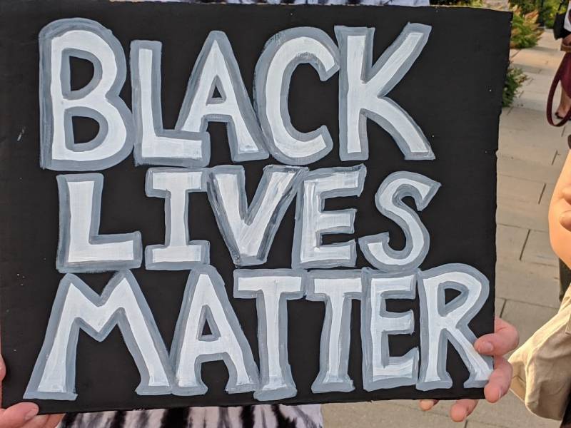 Close-up of a manâ€™s fingers holding a black sign with white lettering highlighted with grey reading â€œBlack Lives Matter,â€ with a part of the manâ€™s black & white shirt and part of a womanâ€™s arm and bag in the background, with sidewalk and planter vegetation in the distance. Photo by Kelley Wegeng.