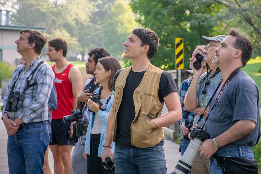 Brody Dunn, President of Champaign County Audubon Society is pictured in the center of a crowd that is looking upward and to the left of the image. Dunn is wearing a brown vest, and holds binoculars in his right hand. Photo by Jen Redwood. 