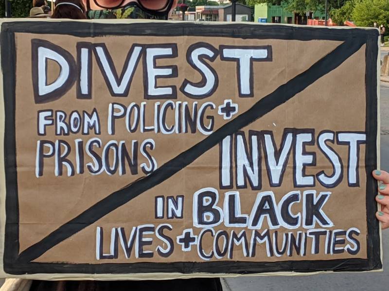 A womanâ€™s hands with seafoam nail polish holding a cardboard sign on which it is painted with a black border and slash reading â€œDivest from Policing + Prisonsâ€ in the upper left triangle and â€œInvest in Black LIves + Communitiesâ€ in the lower right triangle. Peering over the sign is a glimpse of a green and blue masked face wearing black glasses and a streetview in the distance with another bystanderâ€™s hat, some shops, adn a dumpster, as well as two white-masked protesters in the distance. Photo by Kelley Wegeng.