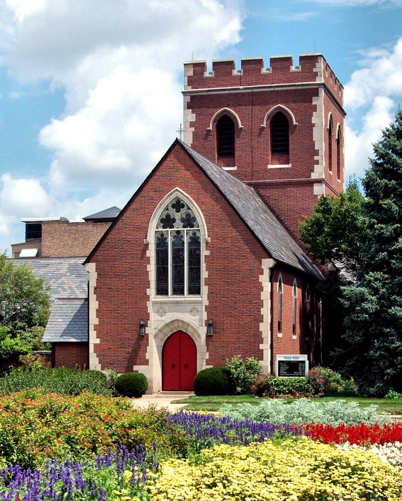 Emmanuel Memorial Episcopal Church, taken facing east from across State Street. Red brick with white stone trim, English Gothic design with rear tower behind gabled narthex. Brightly colored flowers in foreground. Photo by Rick D. Williams.