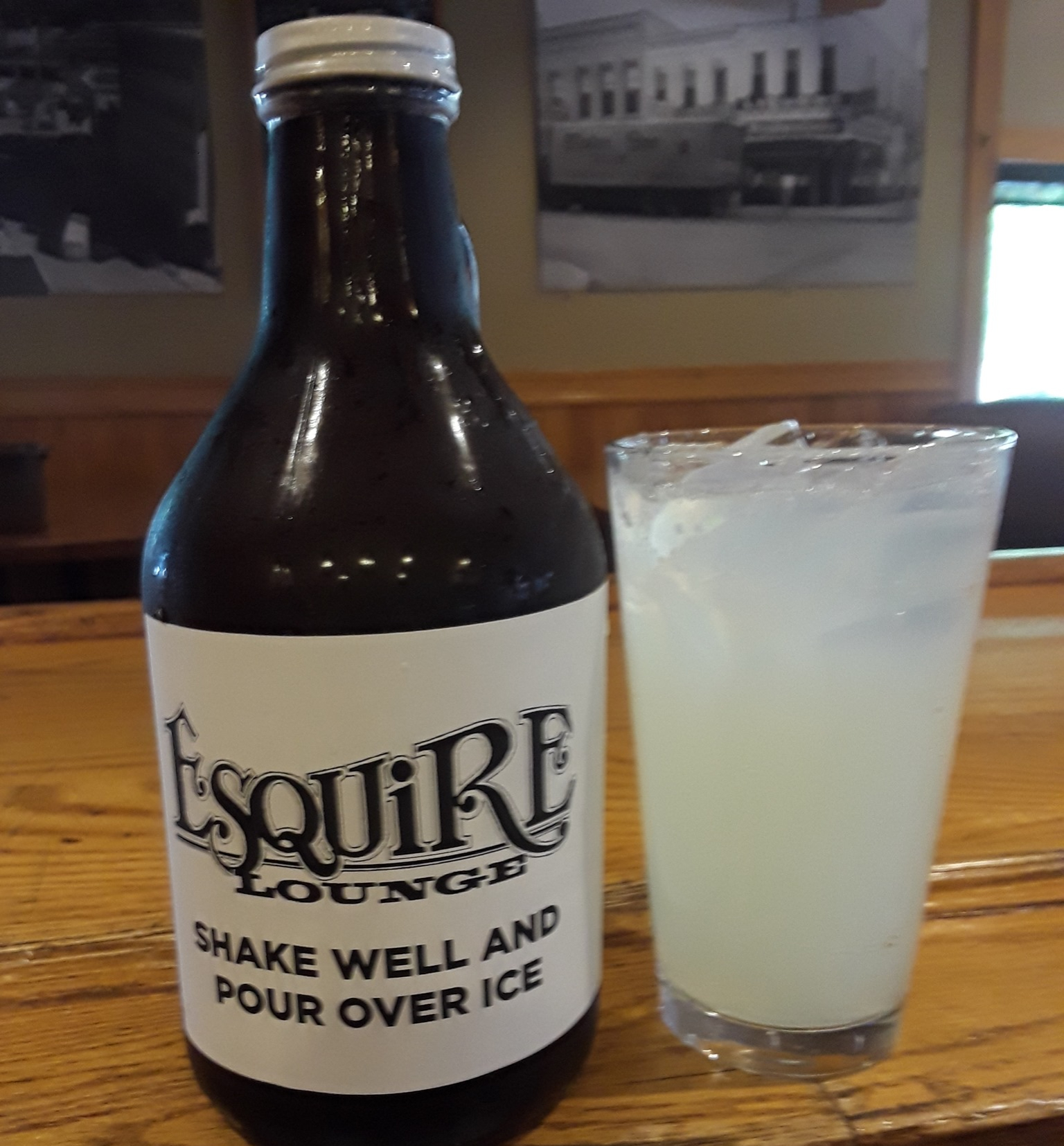 A glass growler sits next to a large glass of a light yellow margarita on a wooden bar. The growler is labeled: Esquire Lounge, shake well and pour over ice. Photo from Esquire's Facebook page.