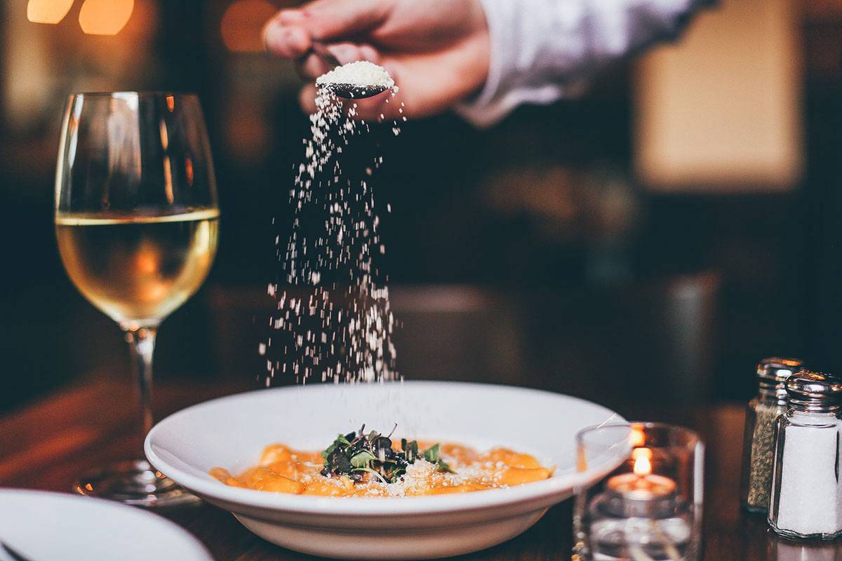 A glass of white wine sits on a table next to a white plate of pasta with parmesan sprinkled on top. Photo from Nando Milano's website.
