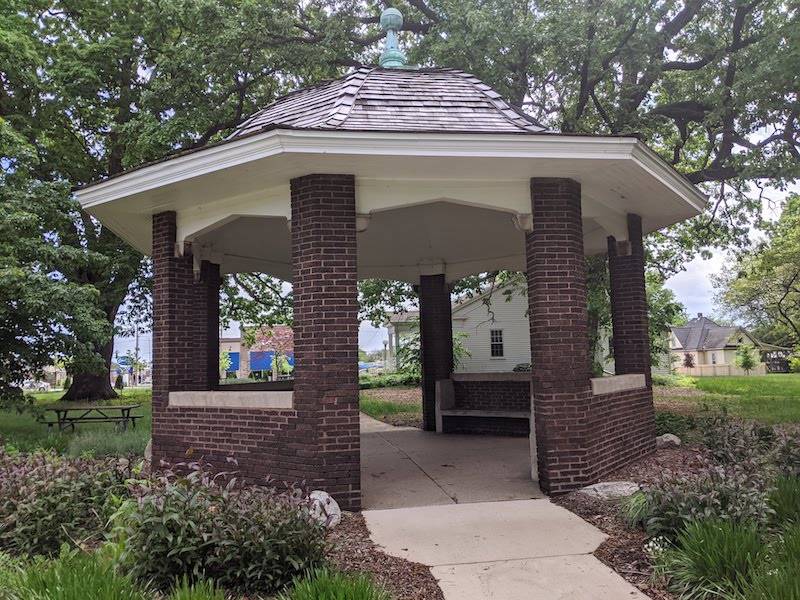 A small gazebo with a red brick base and columns. It has a domed roof with gray shingles, and is surrounded by plants. Photo by Katriena Knights.