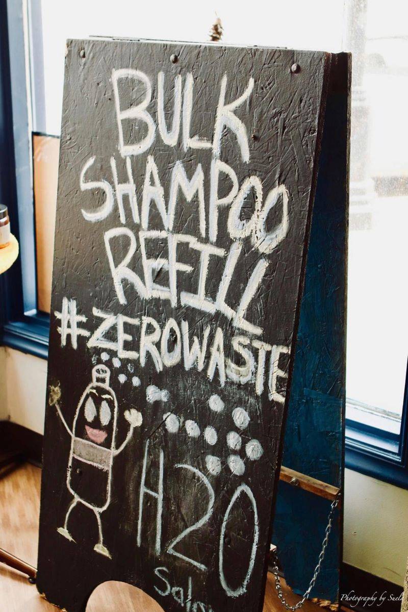A stand up chalk board says Bulk Shampoo Refill #zerowaste, H20 Salon. There is a drawing of a shampoo bottle with arms and legs. Photo by Photography by Suela. 
