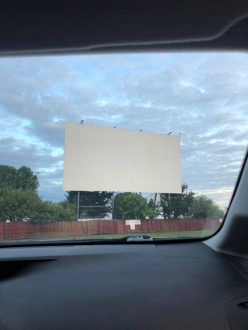 A large outdoor movie screen is visible through the front window of a car. There is a wood fence below it, and a grassy area in the foreground. Photo by Julie McClure.