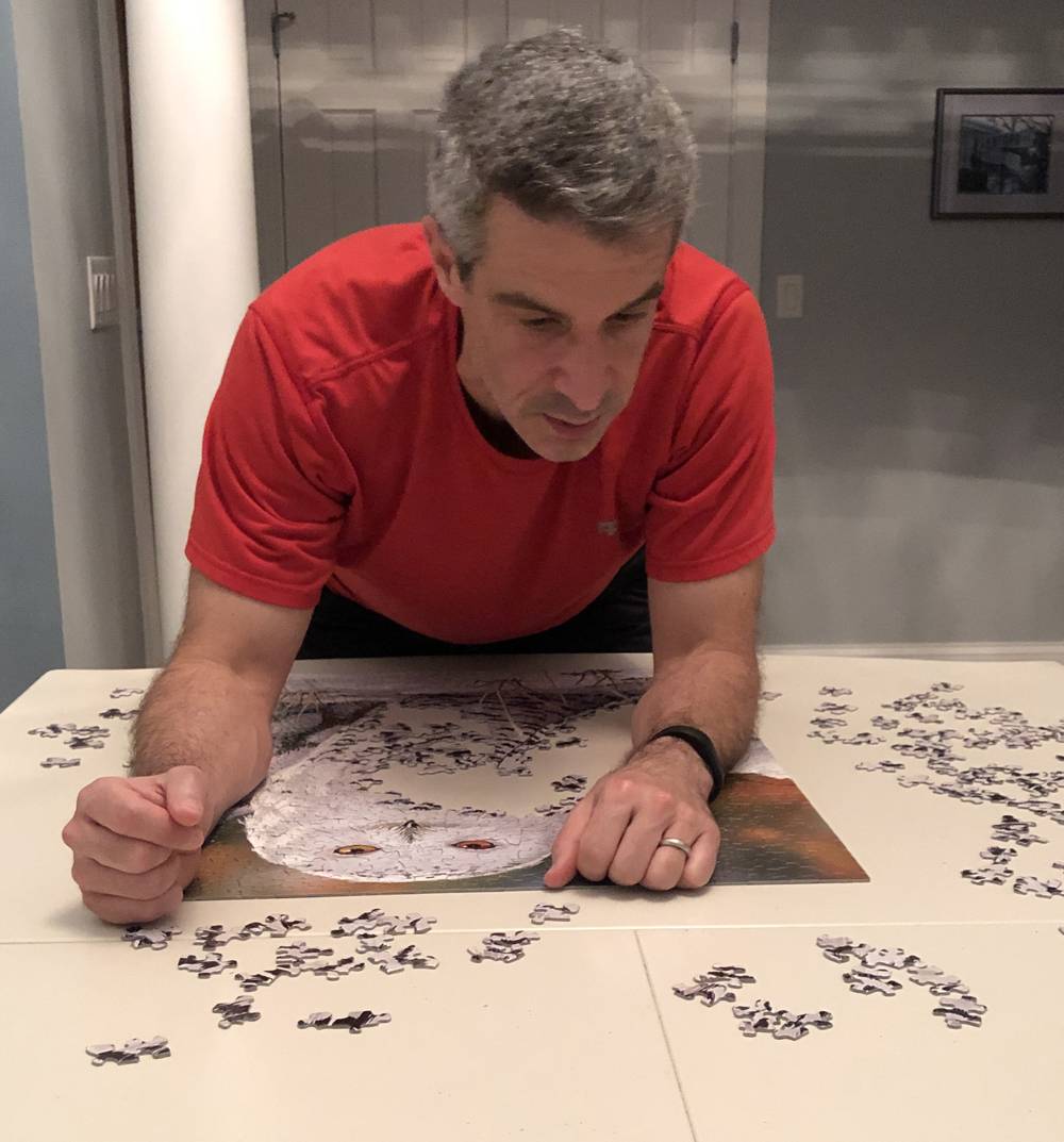 Sanford Hess is wearing a red shirt and is leaning over a table with a mostly completed puzzle featuring an owl. Photo by Elizabeth Hess. 
