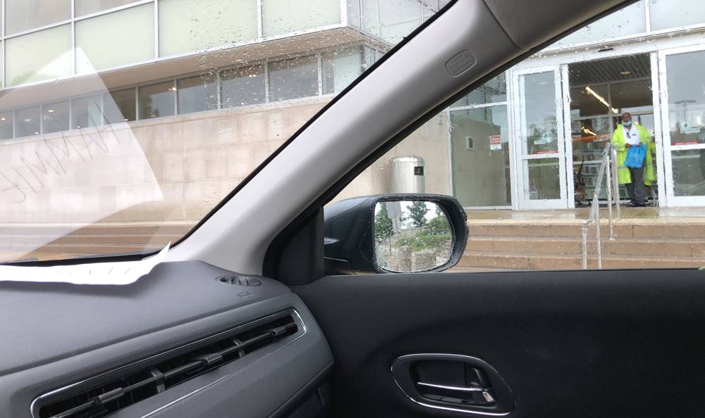 Photo from the driver's seat of a car. On the left is a sheet of paper on the dashboard. There is a name written on it, and it is reflected in the windshield. Through the passenger side window is the Champaign Public Library entrance, with an employee wearing a raincoat in the doorway. Photo by Jessica Hammie. 