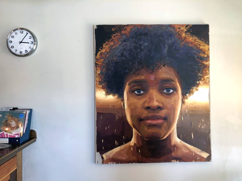 A painting by Patrick Earl Hammie: Ingrid Silva, 2018. The painted portrait is large and done in earth tones and browns. The woman is pictured facing the viewer, unsmiling. She has an Afro. The painting is hung on an off white wall. To the left of the painting is a countertop with a stack of records for sale. Above the records is a wall-hung clock. Photo by Jessica Hammie. 