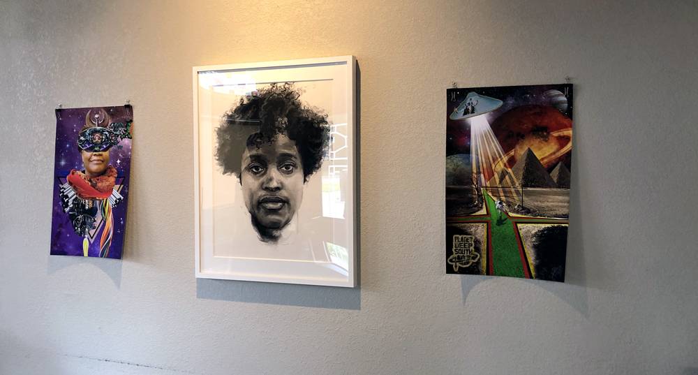 Three vertial artworks are hung on a wall. On the left and on the right are two digital prints with Afrofuturist themes by Stacey Robinson. The left images features a woman's face, though the eyes are obscured by a design. The background is purple and has stars. On the right is an image of a pyramid with a spaceship hovering above and to the left. The spacehship is beaming light down. The background is red. In the center is a portrait of a young black woman with an Afro by Patrick Earl Hammie. Photo by Jessica Hammie. 