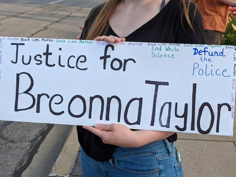 A white sign reading â€œJustice for Breonna Taylorâ€ in black, with small blue print reading â€œDefund the Police,â€ small green print reading â€œEnd White Silence,â€ and in very small print border around the top and sides repeating in green, purple, and black â€œBlack Lives Matter.â€ The torso of the woman holding the sign is visible, she is wearing a black shirt and jeans amidst a street and sidewalk backdrop and an orange shirted man in the background on the right near some green vegetation. Photo by Kelley Wegeng.