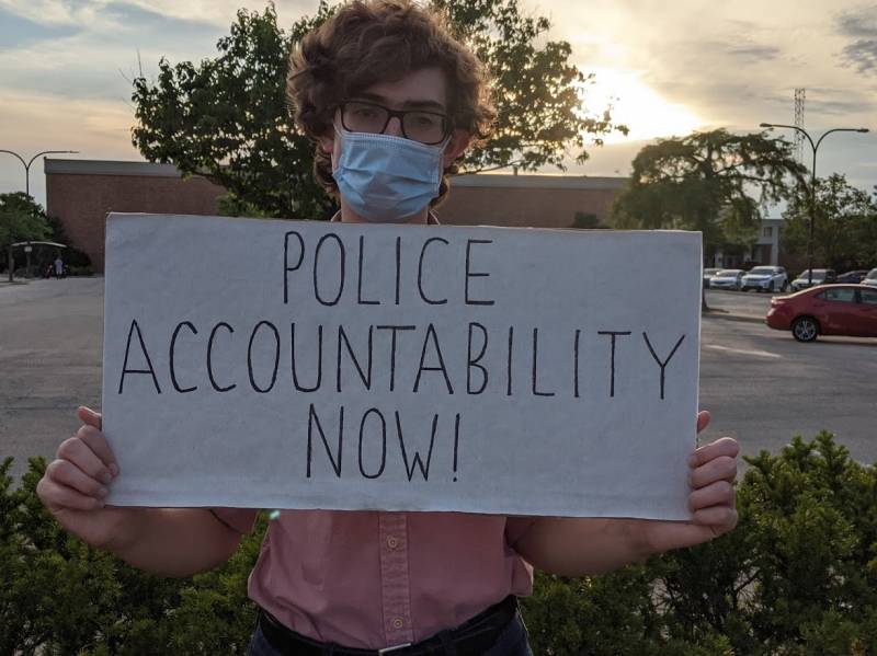 A photo of a young white man with bushy brown hair wearing black glasses, a bluish surgical mask, and pink collared short-sleeved oxford holding a white cardboard sign that reads â€œPOLICE ACCOUNTABILITY NOW!â€ in front of some green parking lot vegetation, parked cars, and the Common Ground Food Coop side of Lincoln Squareâ€™s orange brick, under a partially cloudy sky lit by the setting sun. Photo by Kelley Wegeng.