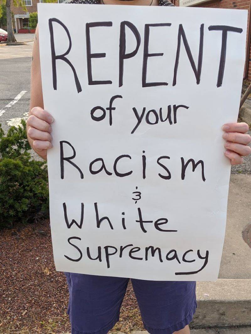 A womanâ€™s hands holding a white sign that says â€œREPENT of your Racism & White Supremacyâ€ in black ink, amidst a background of a parking lot, brick building, and evergreen bush. Her blue shorts are visible below the sign. Photo by Kelley Wegeng.