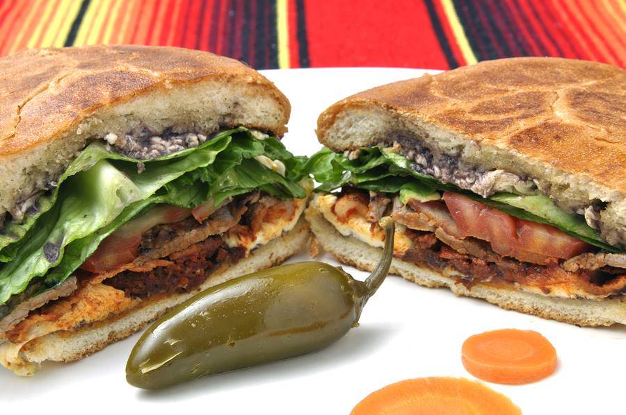  One torta Cubana cut in half displaying the filling of milanesa, hot dog, ham, pinto beans, cheese, lettuce, tomato, onions, avocado, jalapeÃ±os, and mayonnaise. Photo from Taco Motorizado's Facebook page.