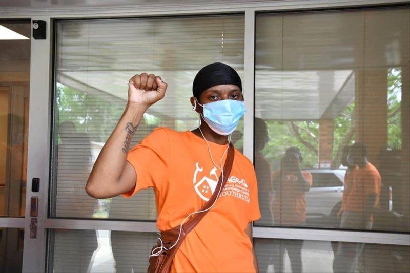 A young adult in an orange t-shirt, mask, and black skull cap is standing with an arm raised and hand in a fist. Photo provided by HACC.