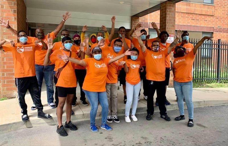 A group of YouthBuild students and leaders, all in masks and orange t-shirts, stand in a group with both arms raised. Photo provided by HACC.