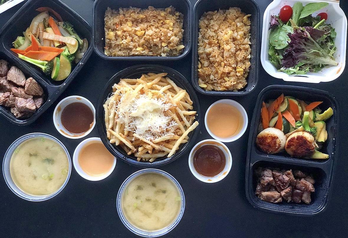 An overhead shot of many dishes. There are two miso soups in circular plastic containers, two side cups of dark brown sauce, two small cups of a light orange sauce, and a small side salad. In the center, there is a black plastic container holding skinny fresh fries with shaved parmesan on top. One divided black container has filet mignon on one side and hibachi vegetables on the other. The other black divided container has steak on one side with two large scallops on top of hibachi vegetables on the other. Photo by Alyssa Buckley.
