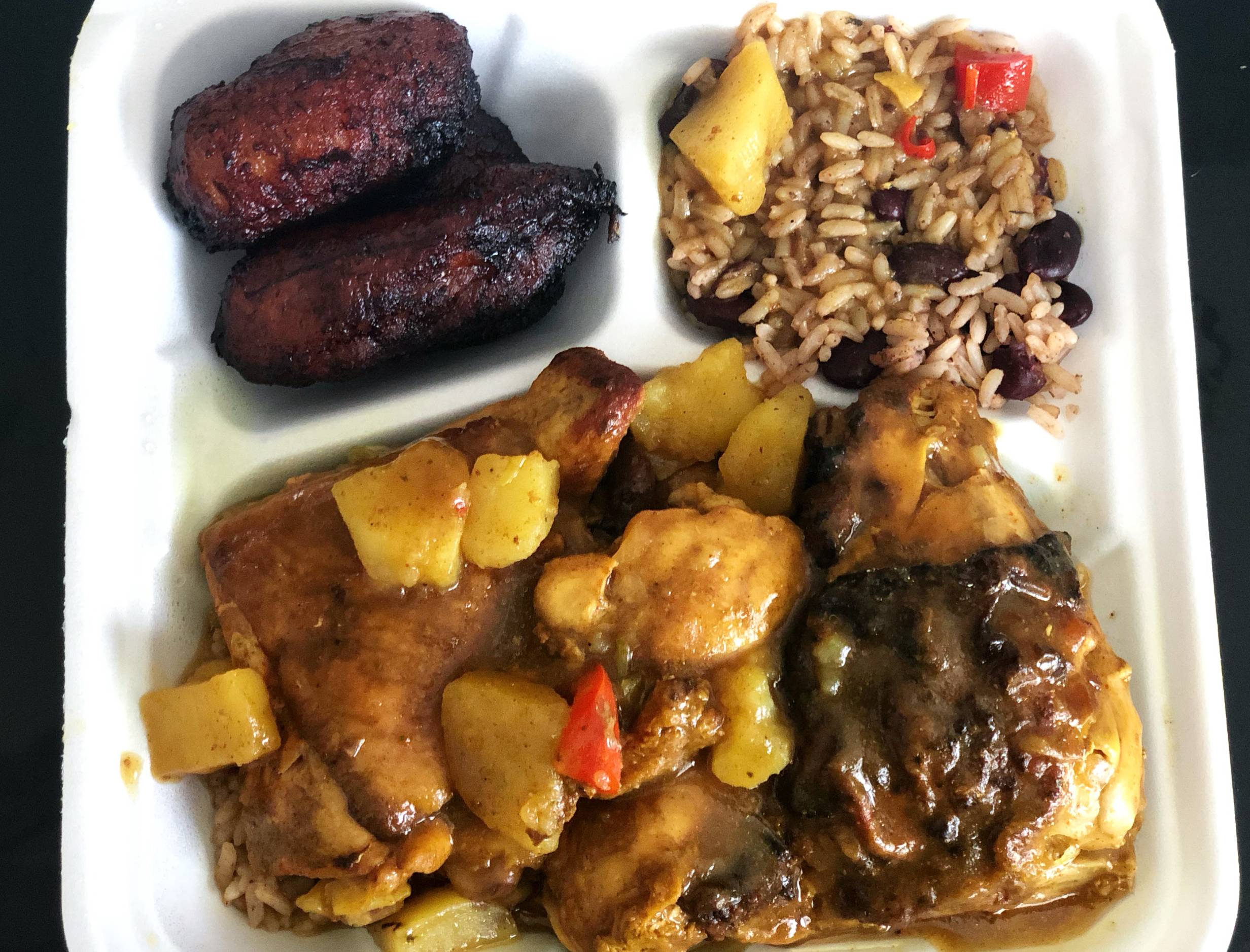 In a white styrofoam divider container, there is a chicken curry with large chunks of potato over brown chicken with a yellow sauce. In the top left divider, there are very dark brown fried plantains. In the top right divider, there are light brown rice with dark red kidney beans. Photo by Alyssa Buckley.