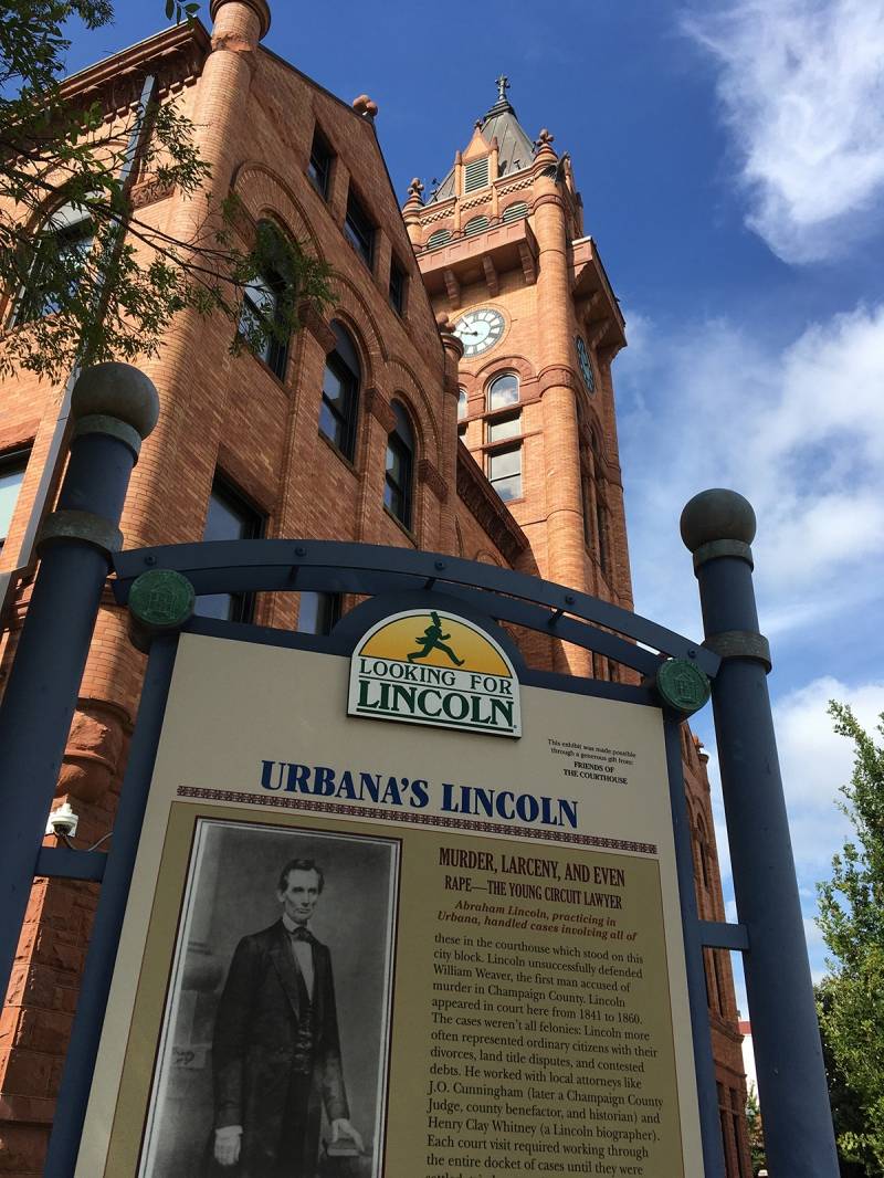 A sign with the heading Urbana's Lincoln stands in front of the red brick courthouse building and clock tower. Photo by Rick D. Williams.