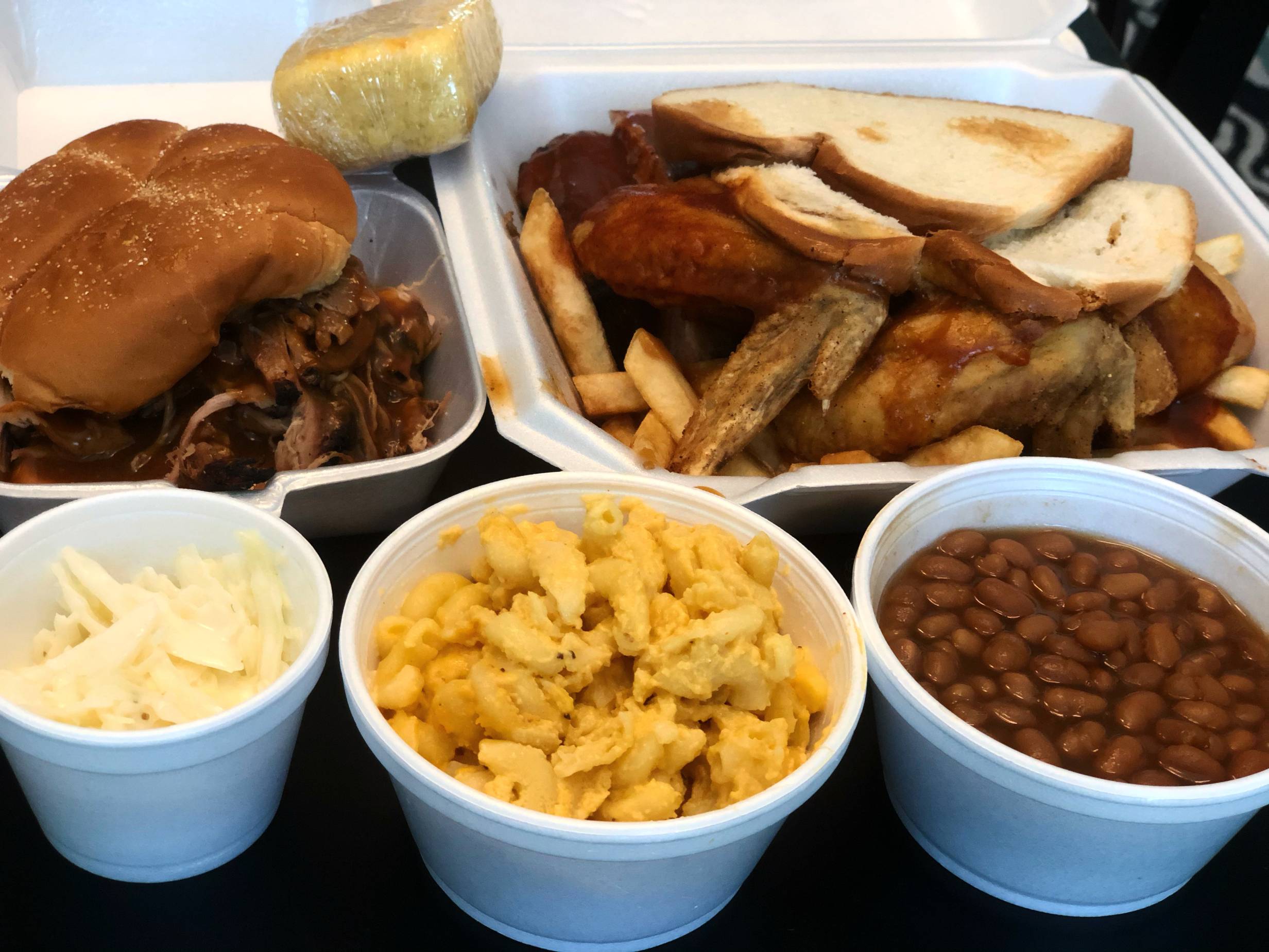 Several styrofoam containers sit open on a black table with a pulled pork sandwich, a ribs and wings combo, coleslaw, macaroni, and baked beans. Photo by Alyssa Buckley.