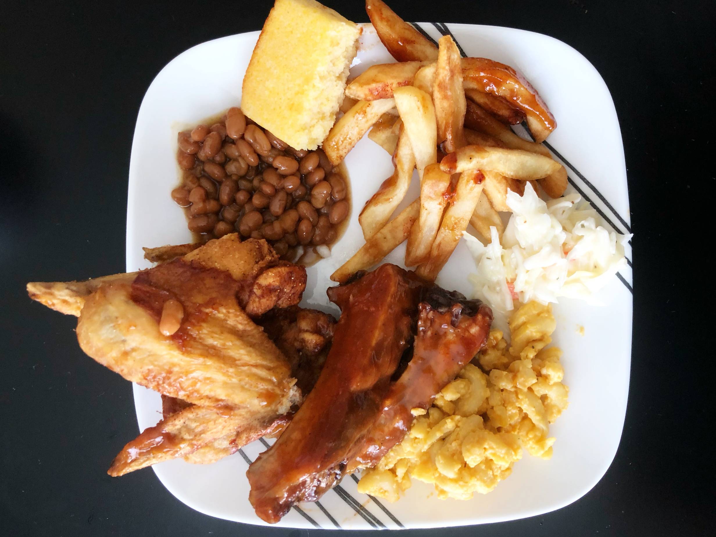 The author's plate from overhead showing wings, beans, cornbread, coleslaw, macaroni, and ribs on a white plate on a black table. Photo by Alyssa Buckley.
