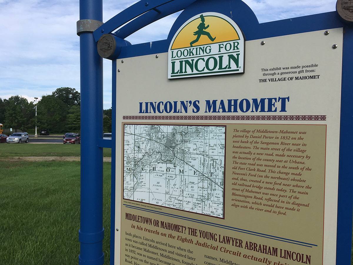 A close up of a sign with the heading Lincoln's Mahomet is in the foreground. In the background there is a grassy field and parking lot. Photo by Rick D. Williams. 