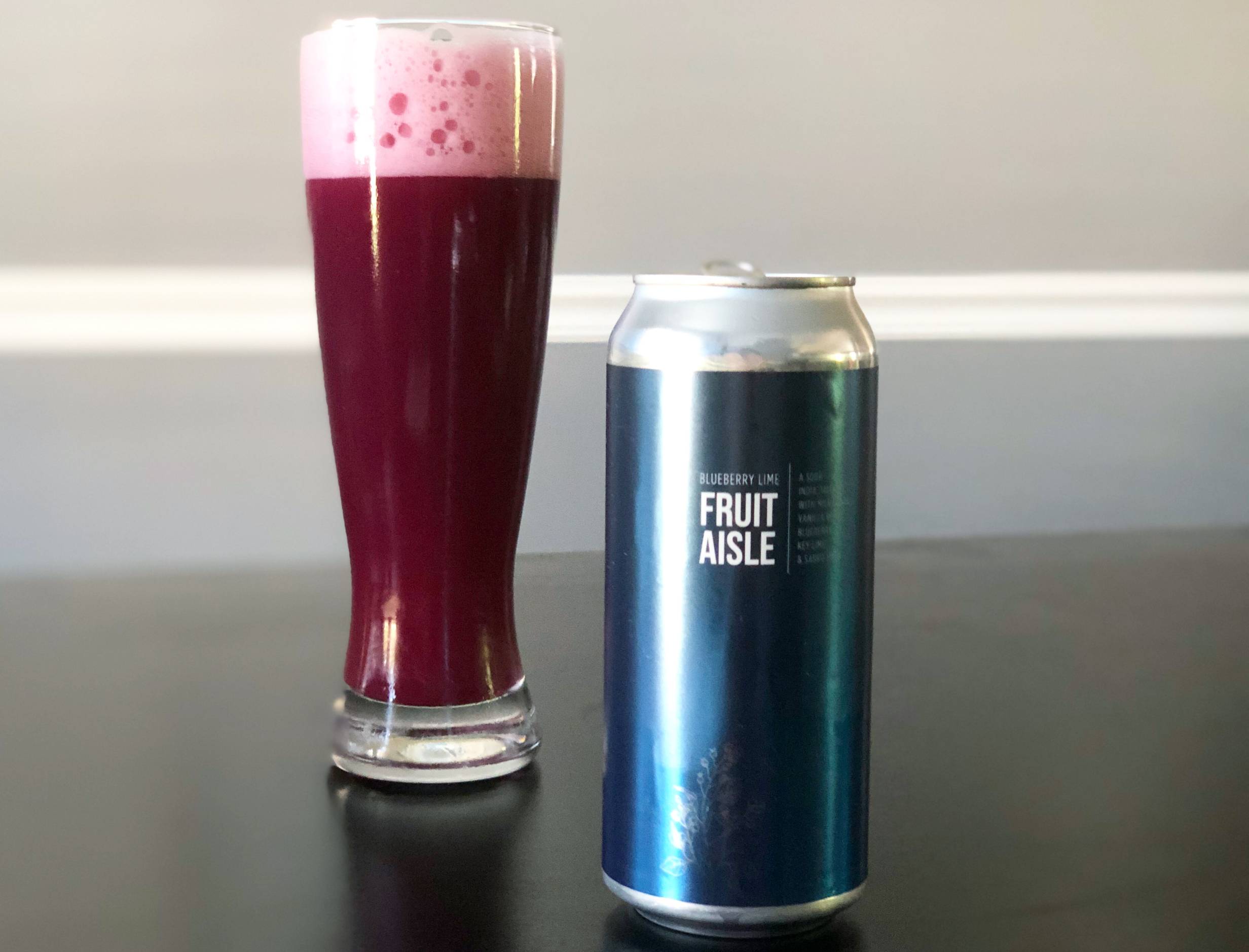 A tall beer glass is full of a pinkish purple beer with an inch of foamy head on a black table next to a dark blue can of Blueberry Lime IPA. Photo by Alyssa Buckley.