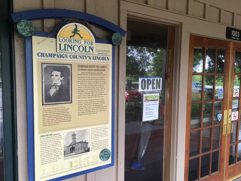 A sign with the heading Champaign County's Lincoln hangs on the outside of a building, alongside a rectangular window and double doors. Photo by Rick D. Williams.