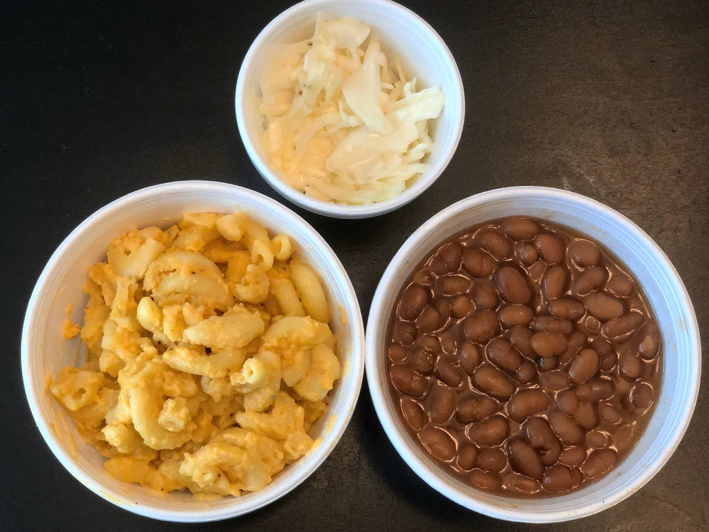 Three white styrofoam containers are open and shot from overhead: a small coleslaw with white cabbage is up top, macaroni on the left, and baked beans on the right. Photo by Alyssa Buckley.