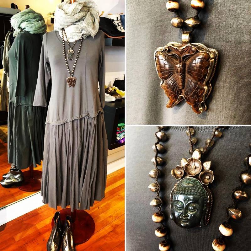 Three images: a full length shot of a mannequin wearing a long gray dress. There is a light gray scarf wrapped around the top and two necklaces with large pendants draped over it. A pair of black boots sit at the bottom. There are close ups of each pendant, one is a butterfly and the other is the face of Buddha. Image from Circles Instagram.