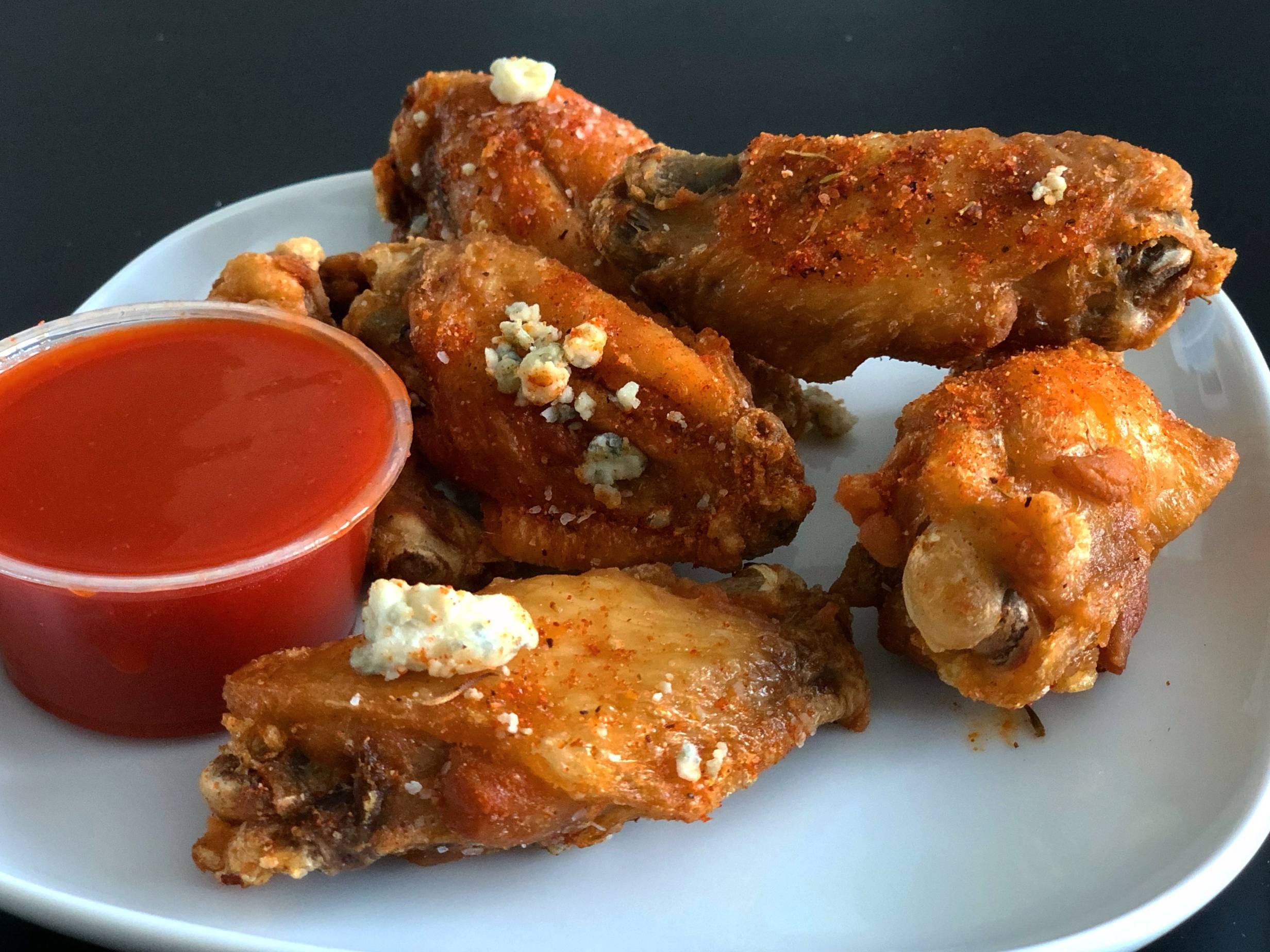 Six fried wings with blue cheese crumbles sit on a white plate with a plastic side cup of bright red dipping sauce. Photo by Alyssa Buckley.