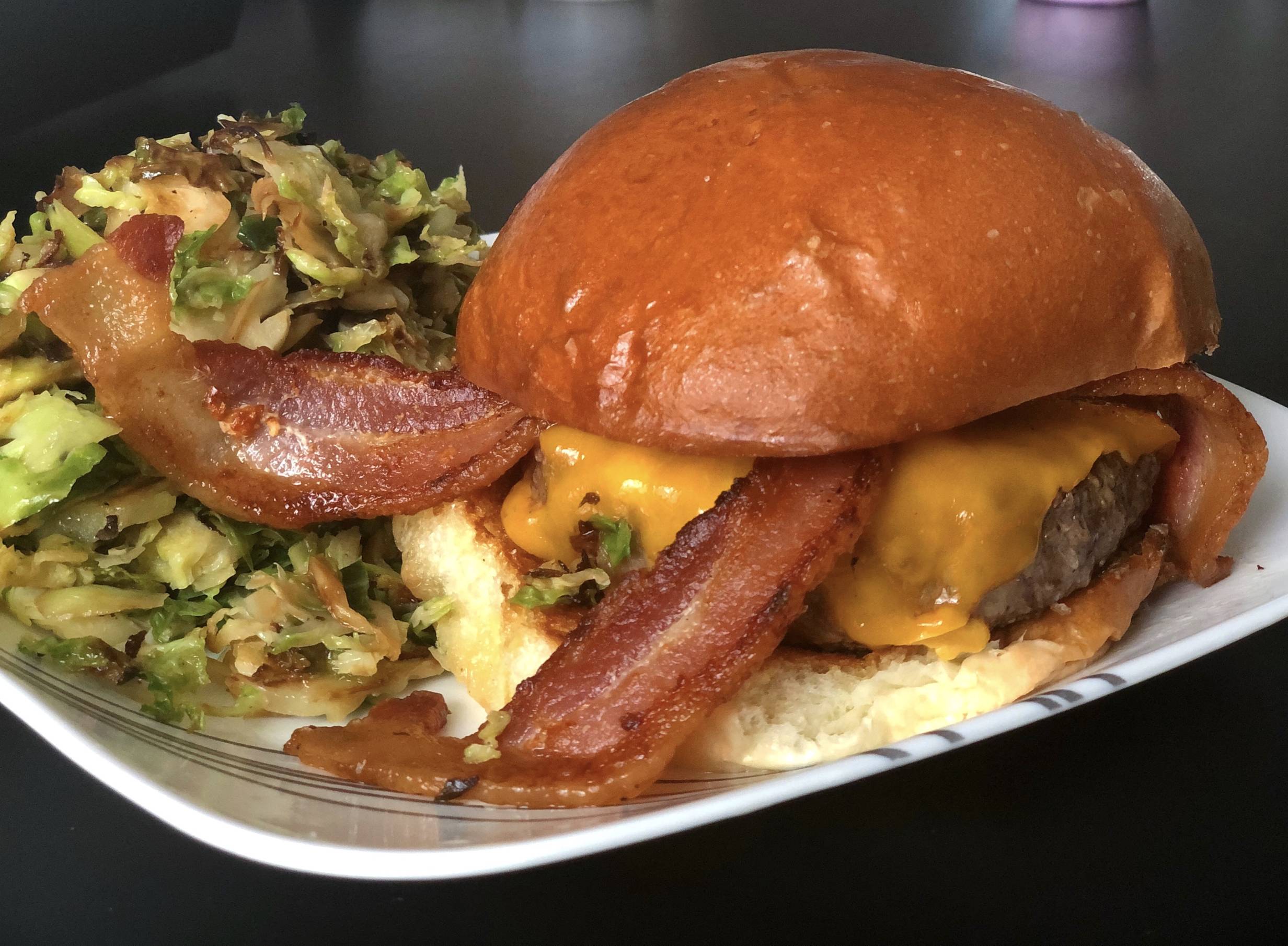 On a white plate, there is a giant cheeseburger with two slices of bacon sticking out several inches next to a pile of shaved Brussels sprouts. Photo by Alyssa Buckley.